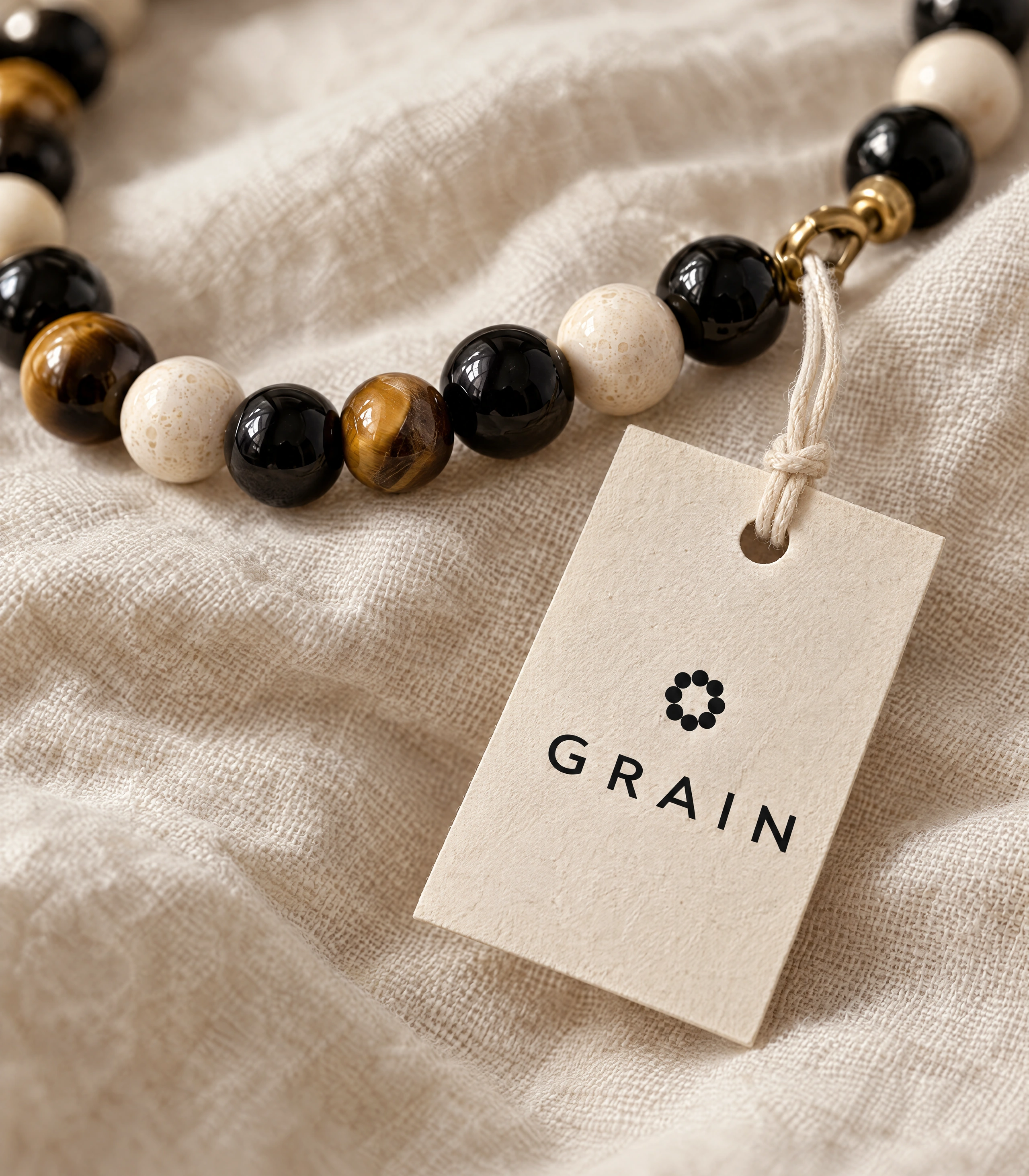Cream GRAIN hang tag tied to a necklace of onyx, tiger's eye and fossil stone on linen