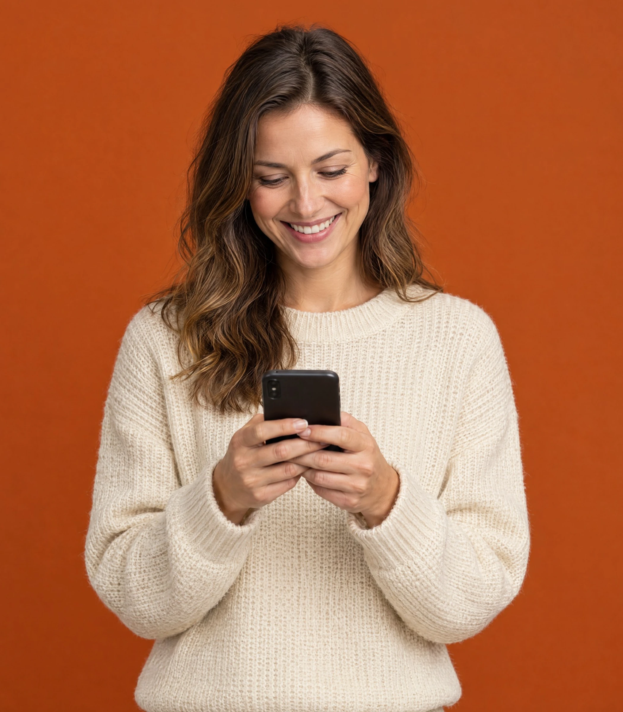 Smiling woman in a cream sweater reading her phone against a burnt-orange wall