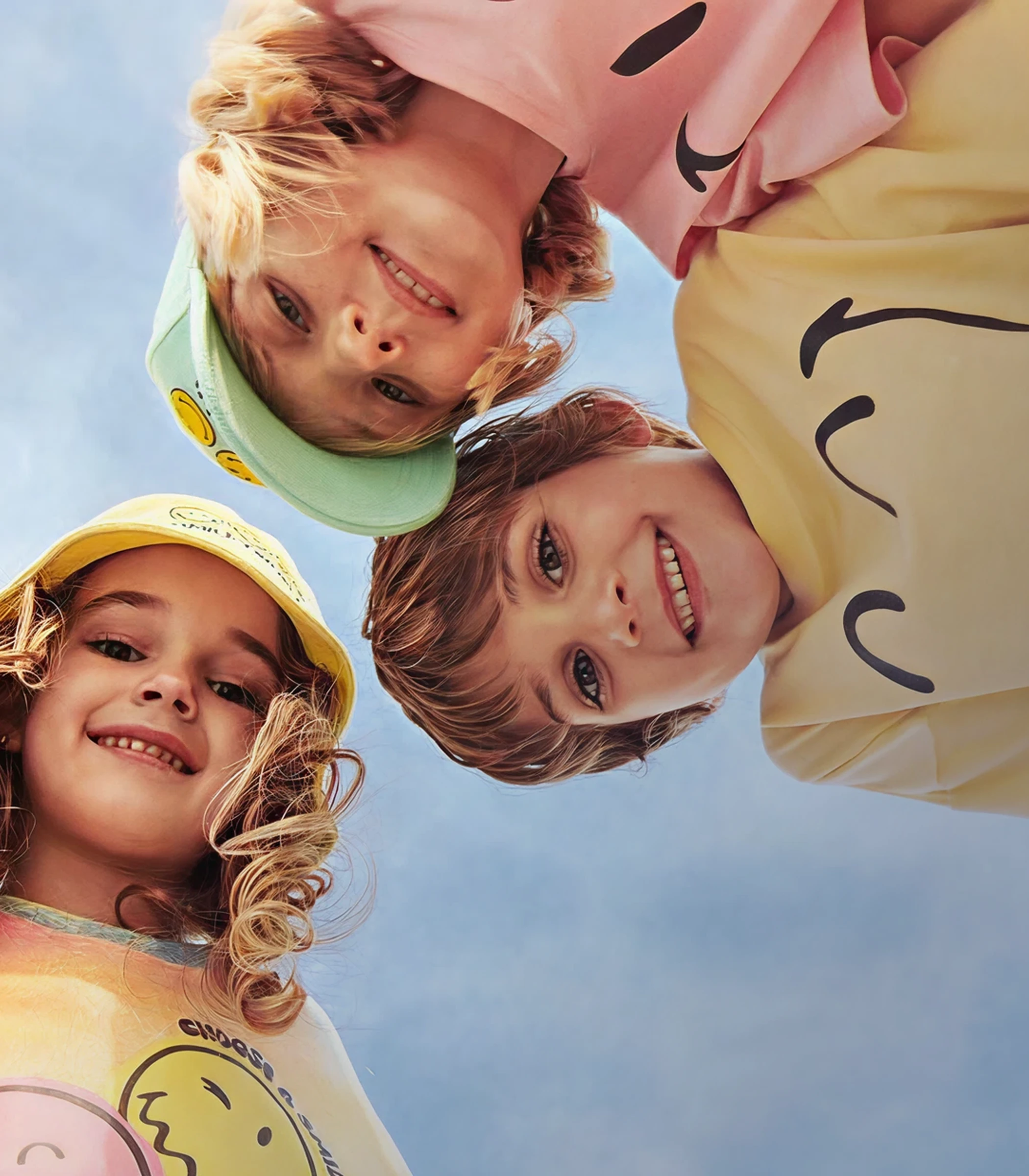 Three smiling children in UNUO clothes looking down into the camera against the sky